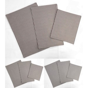 May include: A collection of rectangular fabric placemats. The placemats feature a light gray background with vertical stripes in a darker red-brown color. The placemats are arranged in a layered display, showcasing the striped pattern.