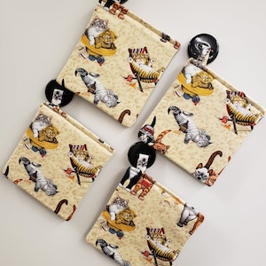 May include: Four beige fabric pouches with a cat design. Each pouch has a black button closure. The cats are depicted in various poses, including lounging in beach chairs and wearing sunglasses.