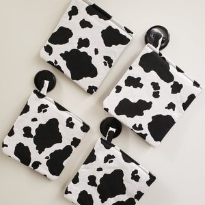 May include: Four square pot holders with a black and white cow print pattern. Each pot holder has a white loop for hanging and is attached to a black hook on a white surface. The pot holders are made of fabric.