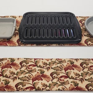 May include: Three baking dishes on a floral patterned kitchen runner. The dishes are a rectangular baking pan, a grill pan, and a round pie pan. All three dishes are made of metal and have a silver finish.