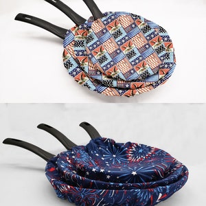 May include: Three black frying pans with red, white, and blue fabric covers. The covers feature a patriotic design with stars, stripes, and eagles. The other set of pans has a blue fabric cover with a firework design.