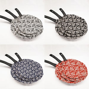 May include: Three black-handled frying pans with patterned fabric covers. The covers are white with black swirls, black with white swirls, and red with white swirls.