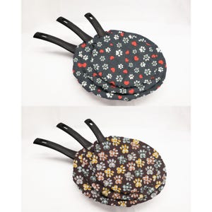 May include: Three black frying pans with paw print and heart patterned fabric covers. The covers are in shades of gray, red, and white, and brown, orange, and yellow.