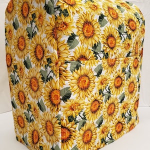 May include: A white kitchen appliance cover with a sunflower pattern. The cover is made of fabric and has a pocket on the side.