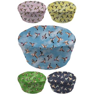 May include: Five fabric hats with hummingbird patterns. The hats are in various colors: light pink, yellow, light blue, green, and navy blue. The hummingbird designs are in flight, with wings spread.