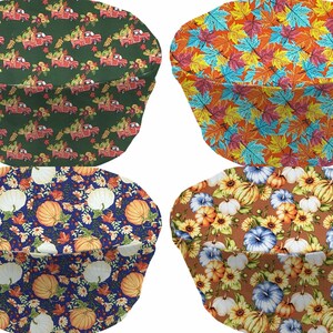 May include: Four round chair cushions with different autumn-themed patterns. The first cushion has a green background with red trucks carrying pumpkins and fall foliage. The second cushion has an orange background with colorful fall leaves. The third cushion has a blue background with white pumpkins, orange pumpkins, and fall leaves. The fourth cushion has a brown background with white pumpkins, blue flowers, and orange pumpkins.