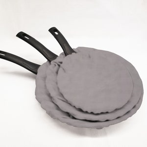 May include: Three black-handled frying pans with gray fabric covers. The pans are stacked on top of each other.