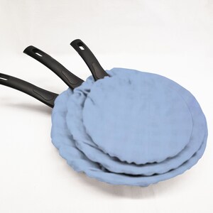 May include: Three black-handled frying pans with blue fabric covers. The pans are stacked on top of each other.
