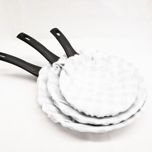 May include: Three black-handled frying pans with white nonstick coating. The pans are stacked on top of each other.