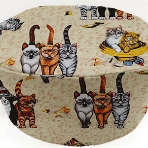 May include: A beige fabric with a repeating pattern of cats wearing sunglasses and hats. The cats are in various poses, including sitting, standing, and lying down. Some of the cats are wearing hats, while others are wearing sunglasses. The fabric is a light beige color with a subtle texture.