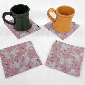 May include: Two mugs, one green and one yellow, are sitting on pink coasters with a bunny rabbit pattern and the text "Happy Easter".