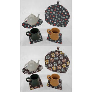 May include: Two sets of teacups and teapot with matching paw print and heart print coasters and tea cozy. The teacups and teapot are white with a glossy finish. The coasters and tea cozy are made of fabric with a dark background and colorful paw prints and hearts.
