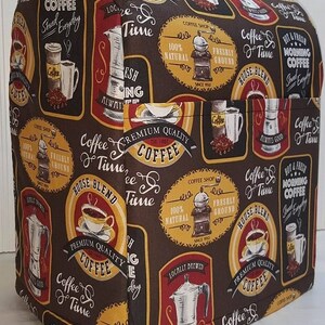 May include: A brown and yellow patterned fabric cover for a kitchen appliance. The cover features a coffee theme with images of coffee cups, coffee beans, and coffee grinders. The text on the cover reads "Coffee Time" and "Premium Quality Coffee".
