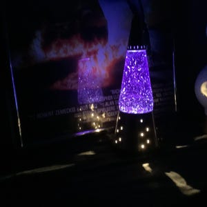 May include: A purple lava lamp with a black base and a conical shape. The lamp is illuminated, with a glowing purple liquid inside. The base has small, round cutouts that emit light. A poster is in the background.
