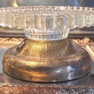 May include: A clear glass pedestal stand featuring a circular top with a beaded edge and a vertical ribbed pattern on the neck. The base is constructed from a tarnished, metallic material with a wide, rounded shape and a visible small hole.