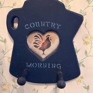 May include: A dark blue wooden wall hook shaped like a coffee pot with a heart-shaped cutout in the center. The cutout features a rooster illustration. The text "COUNTRY" is printed above the heart and "MORNING" below it, with two pegs.