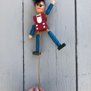 May include: A wooden puppet with blue and red clothing hangs from a string. The puppet has a painted face with a red nose and blue eyes. The puppet is attached to a red ball at the end of the string.
