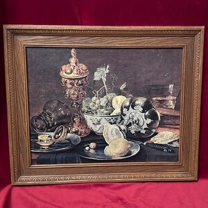May include: A framed still life painting depicting a silver and gold chalice, grapes, a bowl of fruit, and a knife on a table. The painting is set against a dark background.