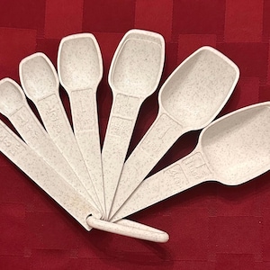 May include: A set of six off-white measuring spoons with a speckled texture, arranged in a fan shape and held together by a plastic ring. Each spoon features embossed measurement markings on the handle, ranging from 1/8 teaspoon to 1 tablespoon.
