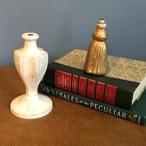 May include: A white ceramic candlestick with a decorative design sits on a brown leather surface next to a stack of books. A gold tassel ornament sits on top of the books. The top book has a green cover with a floral pattern and the text "Tales of the Peculiar" on the spine.