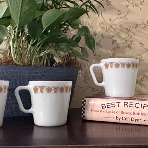 May include: Four white mugs with a gold floral design border. The mugs are arranged on a dark surface with a cookbook titled "Best Recipes" and a potted plant in the background.