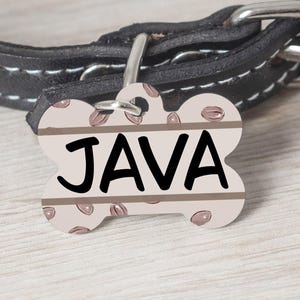 May include: A bone-shaped dog tag with a brown and white coffee bean pattern. The tag has the name "JAVA" printed in black letters.
