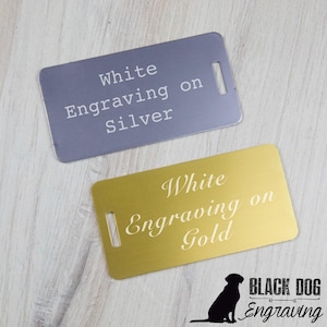 two metal tags with engraved text, one in white and the other in gold, placed on a wooden surface.