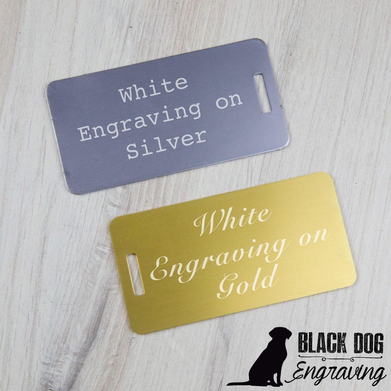 two metal tags with engraved text, one in white and the other in gold, placed on a wooden surface.