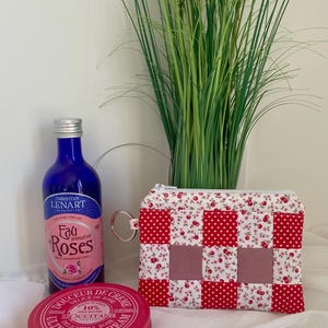 Può includere: Una bottiglia di vetro blu di Eau de Roses Christian Lenart, un barattolo rotondo rosa di crema L'Occitane da 100ml e una pochette rettangolare in tessuto con motivo a scacchi rossi e bianchi, dettagli floreali e una cerniera argentata.