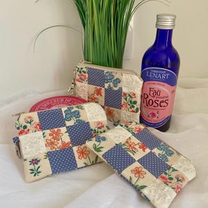Può includere: Una collezione di pochette in tessuto trapuntato e un piccolo beauty case con un motivo floreale e a pois. È visibile una bottiglia di vetro blu di "Eau Roses". Le pochette hanno un design patchwork con accenti crema, blu e floreali.