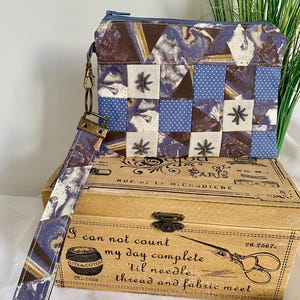 Peut inclure: Une pochette en tissu patchwork avec une dragonne, présentant un mélange de carrés à motifs bleus, marron et blancs. La pochette est posée sur une boîte en bois avec du texte et une illustration de ciseaux.