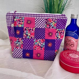 Könnte beinhalten: Eine Kosmetiktasche im Patchwork-Stil in Lila, Rosa und Weiß mit Blumen- und Tupfenmustern. Die Tasche hat einen Reißverschluss mit Quaste. Eine blaue Flasche und eine rote Dose liegen neben der Tasche.