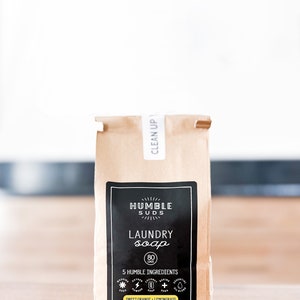 Puede incluir: Una bolsa de papel marr&oacute;n con una etiqueta negra que dice "HUMBLE SUDS LAUNDRY soap 80 LOADS 5 HUMBLE INGREDIENTS SWEET ORANGE - LEMONGRASS FOR ALL MACHINES 40 OZ | THIS BAG IS COMPOSTABLE" y una etiqueta blanca que dice "CLEAN UP".