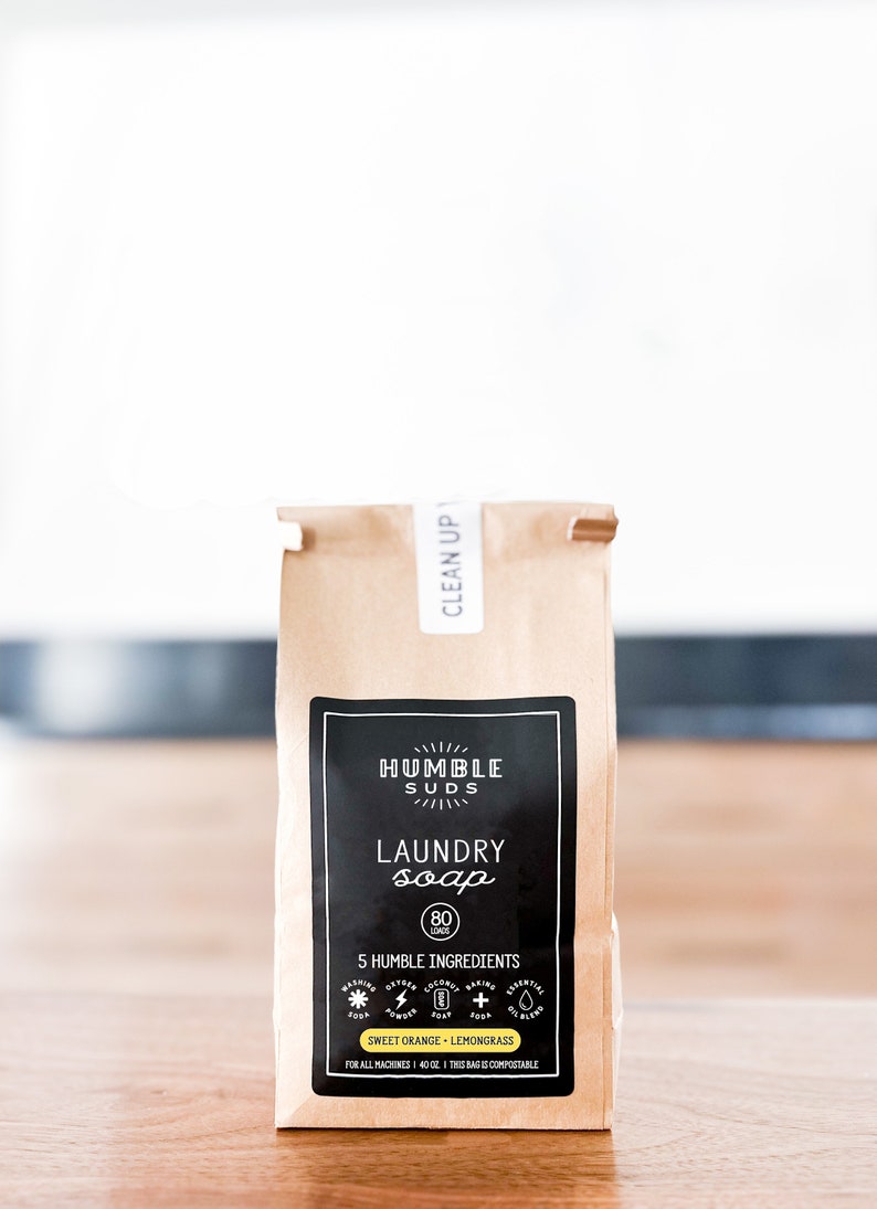 Puede incluir: Una bolsa de papel marr&oacute;n con una etiqueta negra que dice "HUMBLE SUDS LAUNDRY soap 80 LOADS 5 HUMBLE INGREDIENTS SWEET ORANGE - LEMONGRASS FOR ALL MACHINES 40 OZ | THIS BAG IS COMPOSTABLE" y una etiqueta blanca que dice "CLEAN UP".