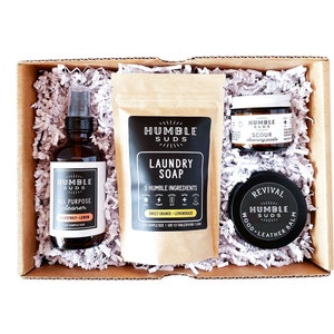 May include: A gift box filled with natural cleaning products from Humble Suds. The box contains a brown paper bag of laundry soap, a glass bottle of all-purpose cleaner, a small jar of cleaning paste, and a small jar of wood and leather balm.