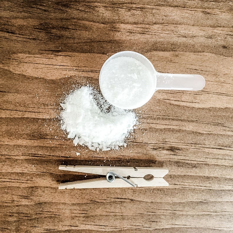 Puede incluir: Una cuchara dosificadora de pl&aacute;stico blanco con polvo blanco que se derrama sobre una superficie de madera. Tambi&eacute;n hay una pinza de madera en la superficie.