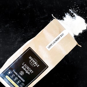 Puede incluir: Una bolsa de papel marr&oacute;n con una etiqueta blanca que dice "HUMBLE SUDS LAUNDRY soap 100 LOADS 5 HUMBLE INGREDIENTS SWEET ORANGE - LEMONGRASS" y una etiqueta blanca m&aacute;s peque&ntilde;a que dice "LOVE LAUNDRY DAY". Polvo de detergente blanco se derrama de la bolsa sobre una superficie negra.