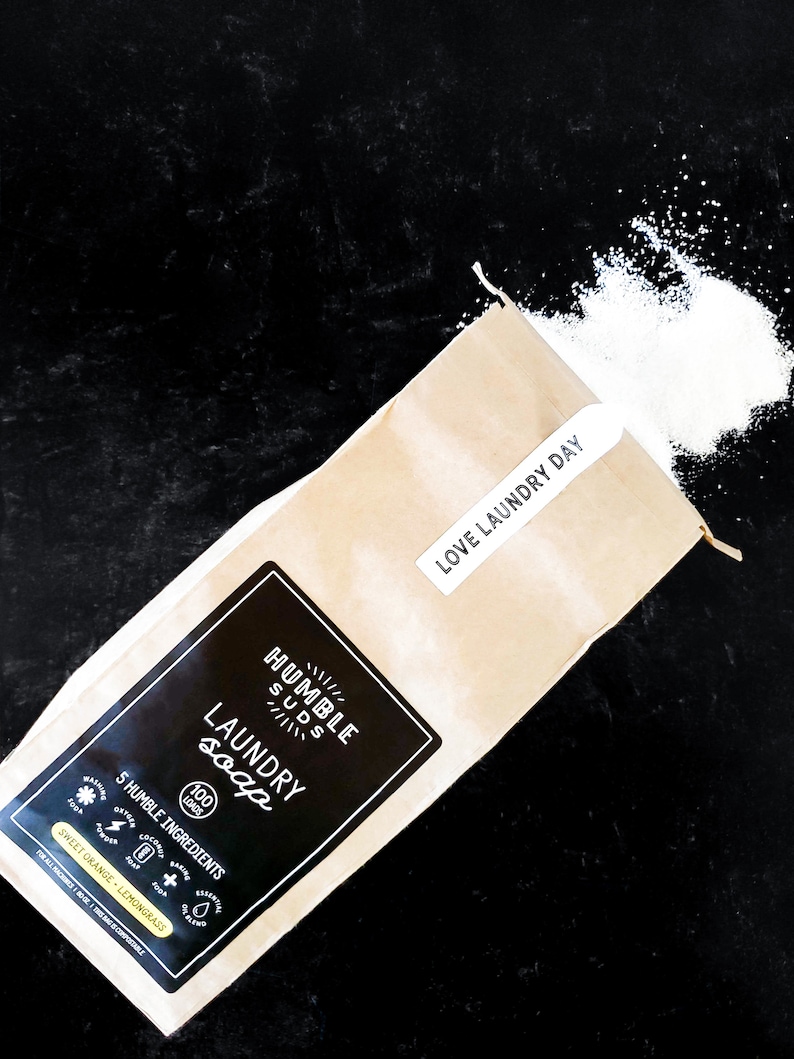 Puede incluir: Una bolsa de papel marr&oacute;n con una etiqueta blanca que dice "HUMBLE SUDS LAUNDRY soap 100 LOADS 5 HUMBLE INGREDIENTS SWEET ORANGE - LEMONGRASS" y una etiqueta blanca m&aacute;s peque&ntilde;a que dice "LOVE LAUNDRY DAY". Polvo de detergente blanco se derrama de la bolsa sobre una superficie negra.