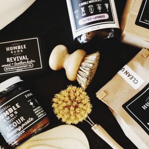 Puede incluir: Una colecci&oacute;n de productos de limpieza naturales de Humble Suds, que incluye un limpiador multiusos, un b&aacute;lsamo para madera y cuero, una pasta para fregar y herramientas de limpieza. Los productos est&aacute;n empaquetados en bolsas de papel marr&oacute;n y tienen etiquetas en blanco y negro con el logotipo de la empresa y los nombres de los productos.