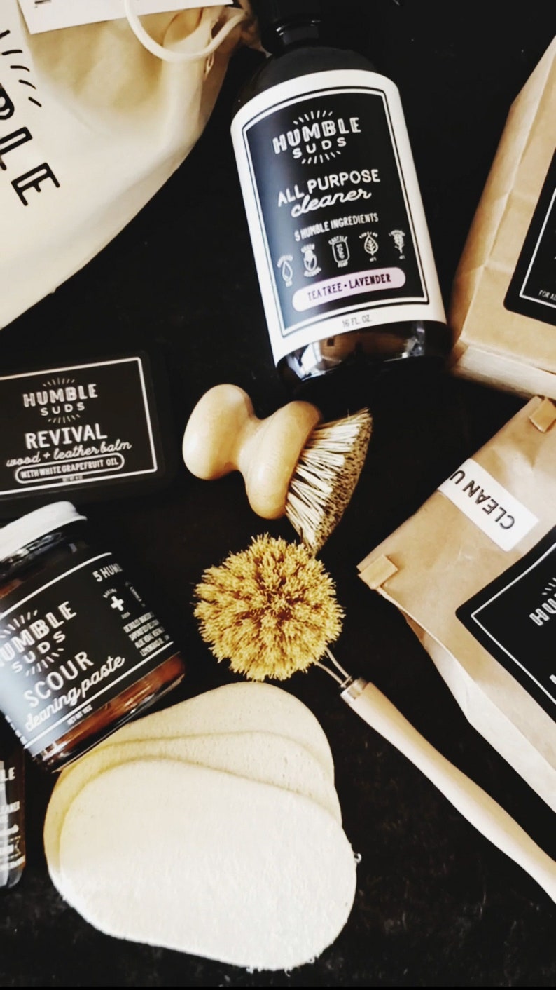 Puede incluir: Una colecci&oacute;n de productos de limpieza naturales de Humble Suds, que incluye un limpiador multiusos, un b&aacute;lsamo para madera y cuero, una pasta para fregar y herramientas de limpieza. Los productos est&aacute;n empaquetados en bolsas de papel marr&oacute;n y tienen etiquetas en blanco y negro con el logotipo de la empresa y los nombres de los productos.