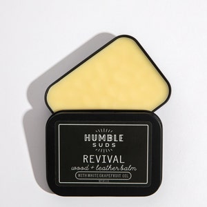 May include: A black tin with a white label that reads "Humble Suds Revival wood + leather balm with white grapefruit oil" and a black lid with a yellow balm inside.