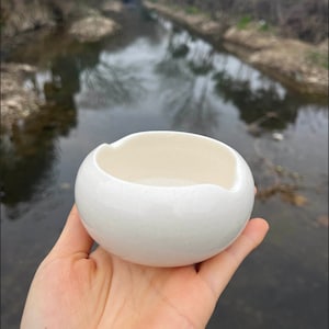 Puede incluir: Un pequeño cuenco de cerámica de color blanco roto, de forma redonda, con un acabado liso y brillante y una textura moteada. El cuenco presenta dos muescas curvas distintas en los lados opuestos del borde, creando un diseño funcional.