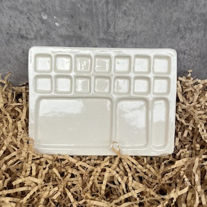 Peut inclure: Une palette d'aquarelle rectangulaire en céramique blanc cassé, dotée de douze petits godets carrés disposés sur deux rangées en haut, ainsi que deux zones de mélange rectangulaires plus grandes situées au bas du plateau.