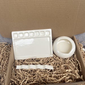 May include: A set of three off-white ceramic art supplies consisting of a rectangular paint palette with six small wells, a round water bowl with a smooth rim, and a long, narrow, wavy brush rest, all nestled inside a cardboard box with paper filler.