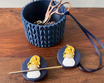 May include: Two blue and white gnome needle minders with a yellow and white gnome on top. The minders are on a wooden surface with a blue container with a pair of scissors and a blue cord.