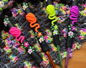 May include: Four colorful crochet hooks with floral and yarn ball embellishments. The hooks are black with a silver metal hook. The yarn balls are pink, green, orange, and purple.