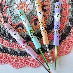 Può includere: Quattro uncinetti con manici color pastello. Ogni manico è decorato con un fantasma bianco e piccoli fiori. Gli uncinetti sono disposti su una tovaglietta all'uncinetto nei toni del rosa, grigio e verde. Per l'uncinetto.