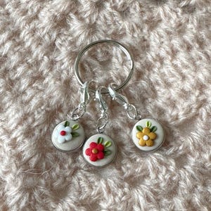 Pode incluir: Um conjunto de três marcadores de ponto florais em um chaveiro prateado. Cada marcador apresenta uma pequena base redonda esbranquiçada com um design floral colorido: branco, vermelho e amarelo. Os marcadores são projetados para tricô ou crochê.
