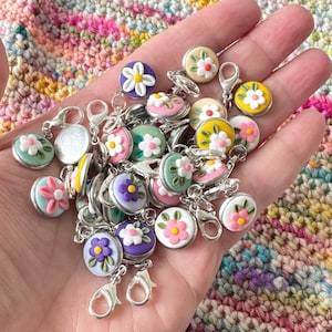May include: A collection of colorful flower-shaped stitch markers with silver clasps. The markers are made of polymer clay and feature various flower designs with white, pink, purple, and yellow accents.