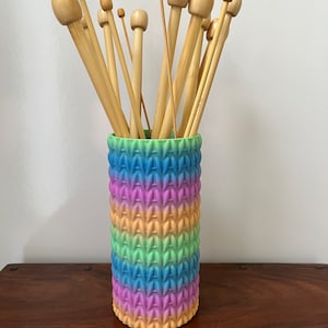 Può includere: Un contenitore cilindrico color arcobaleno con una superficie testurizzata contiene una collezione di ferri da maglia in legno.
